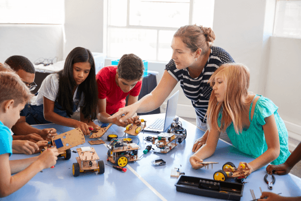 Group of children working on robotics projects at a table while a woman assists and guides them.